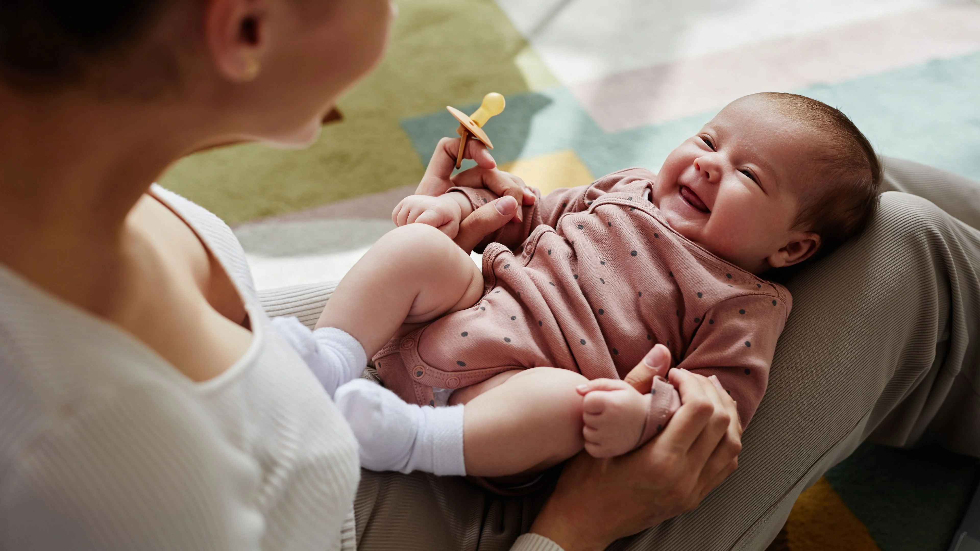 A 10 week old baby lying on a bed smiling up at a parent who is leaning in and talking softly, warm natural light, candid family photograph