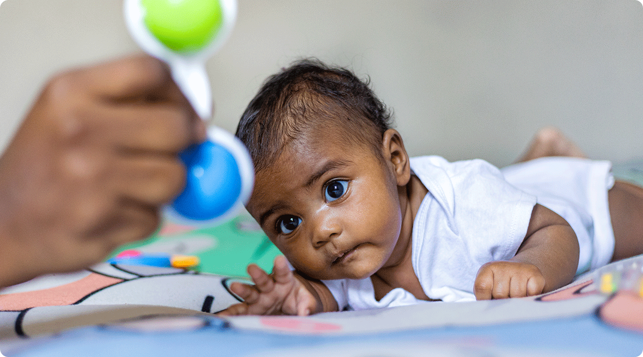 A baby doing tummy time on a clean foam play mat, lifting their head and chest while reaching for a colorful toy, parent sitting nearby with hands ready, warm daylight