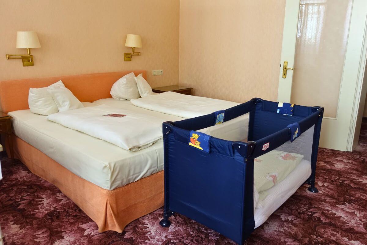 A baby sleeping in a portable travel crib in a dim hotel room with a white noise machine on a dresser, natural candid photo