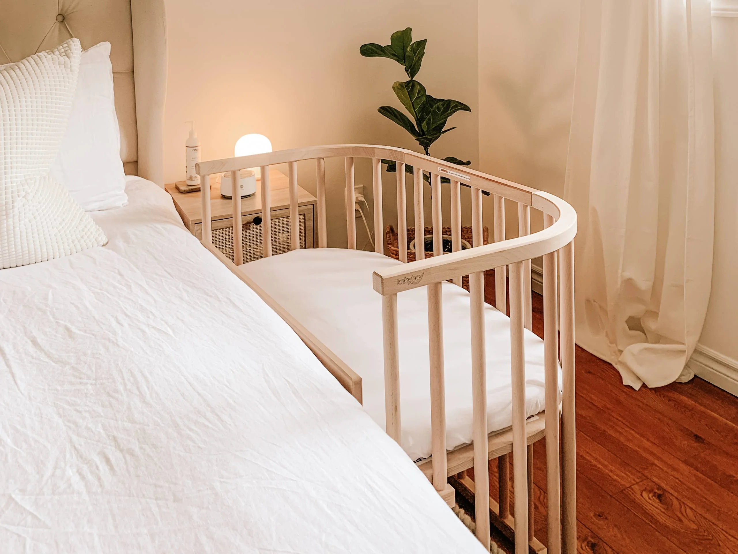 A bedside bassinet positioned next to an adult bed in a dim, calm bedroom at night with a small lamp glowing, photorealistic lifestyle photograph