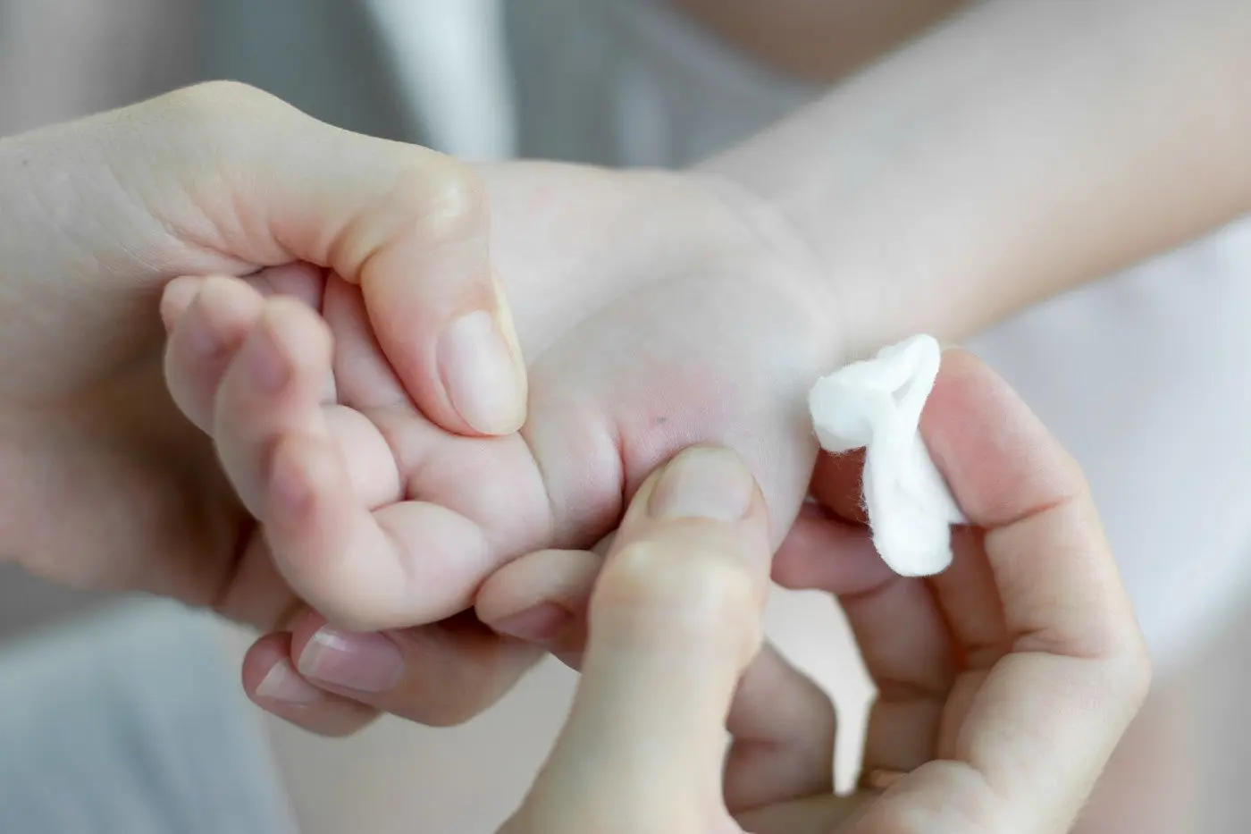A caregiver gently holding a toddler’s hand under warm light while preparing to remove a small splinter with clean tweezers on a nearby counter, real-life photo