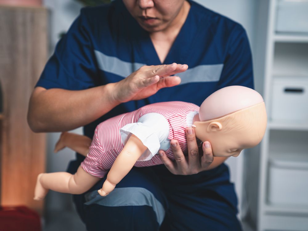 A caregiver giving firm back blows between an infant's shoulder blades while the baby is positioned face-down on the caregiver's forearm, head supported, realistic home setting photo