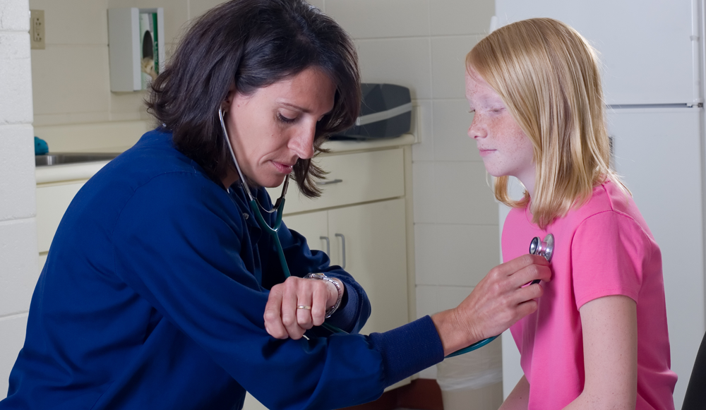 A caregiver sitting at a kitchen table reviewing a child’s allergy action plan paperwork with a daycare provider, with an epinephrine auto-injector trainer on the table, realistic photo