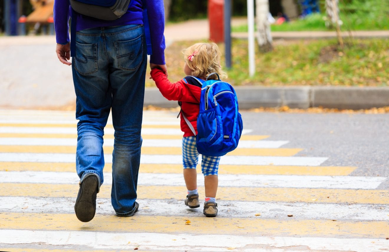 A childcare provider walking outdoors while holding a toddler’s hands gently with the toddler’s arms relaxed, realistic photo