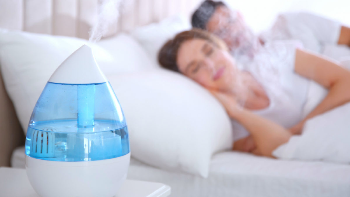 A cool mist humidifier on a dresser in a child bedroom at night with soft lamp light, emitting a visible mist plume, realistic photo
