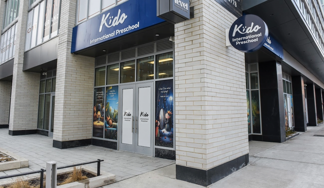 A daycare teacher kneeling at a classroom door with open arms while a parent holds a hesitant toddler, morning drop-off moment, natural light photo