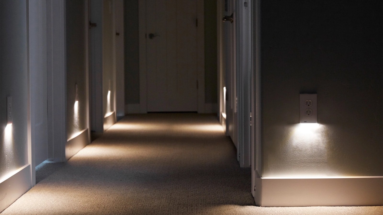 A dimly lit hallway at night with a small plug-in nightlight illuminating the path toward a bathroom door, photorealistic home interior photography