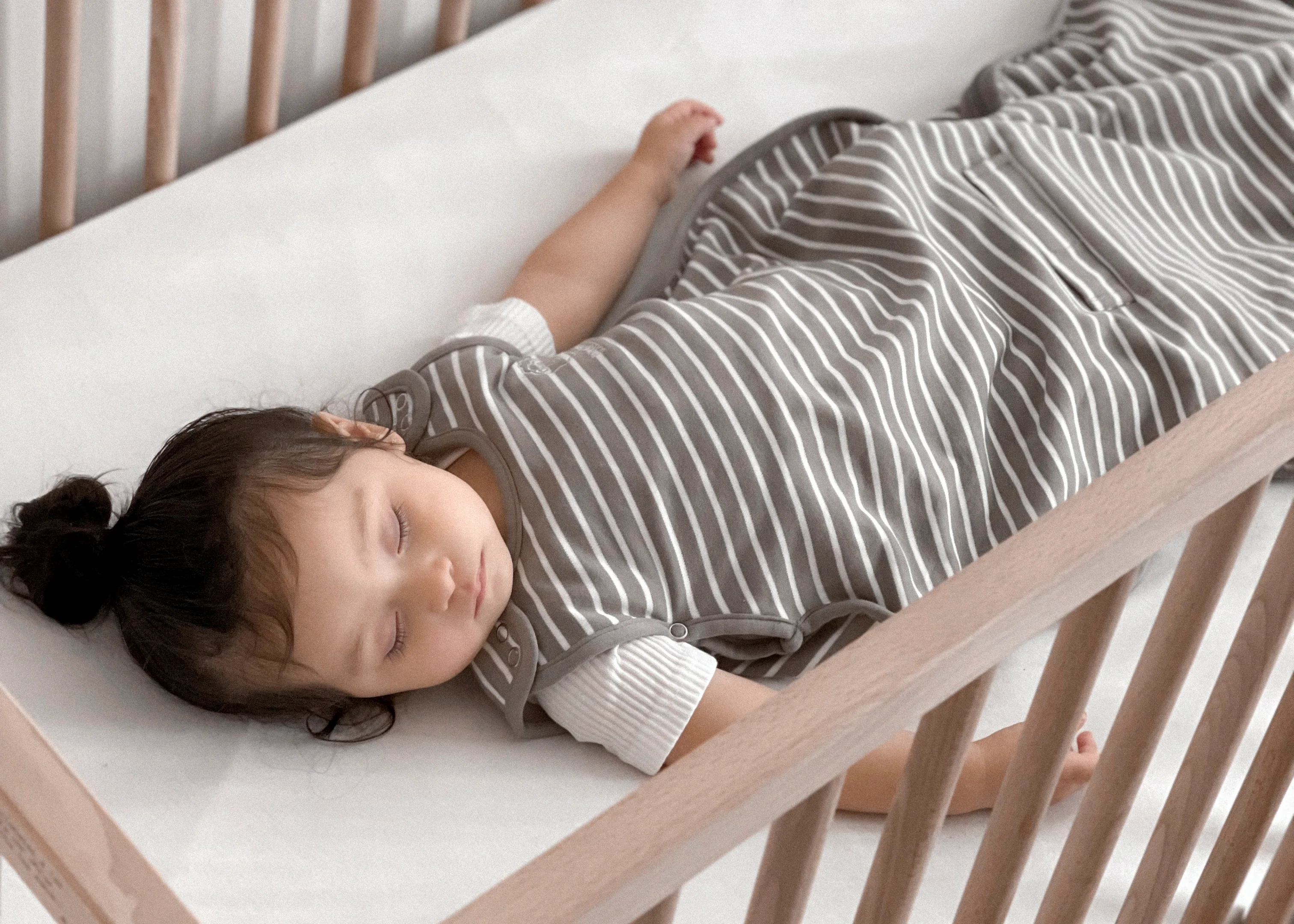 A four-month-old baby sleeping on their back in a crib wearing a sleep sack, soft daylight coming through curtains, calm home photography