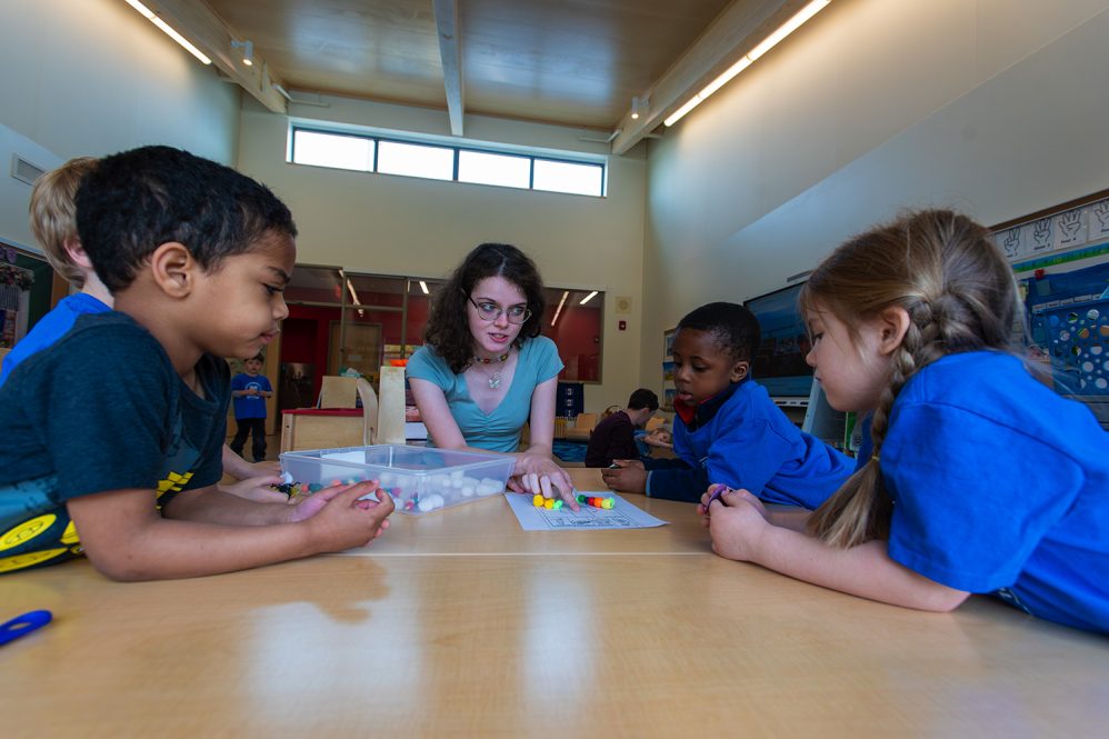 A kindergarten teacher crouching to speak calmly with an upset young child near a classroom reading corner, realistic school photograph