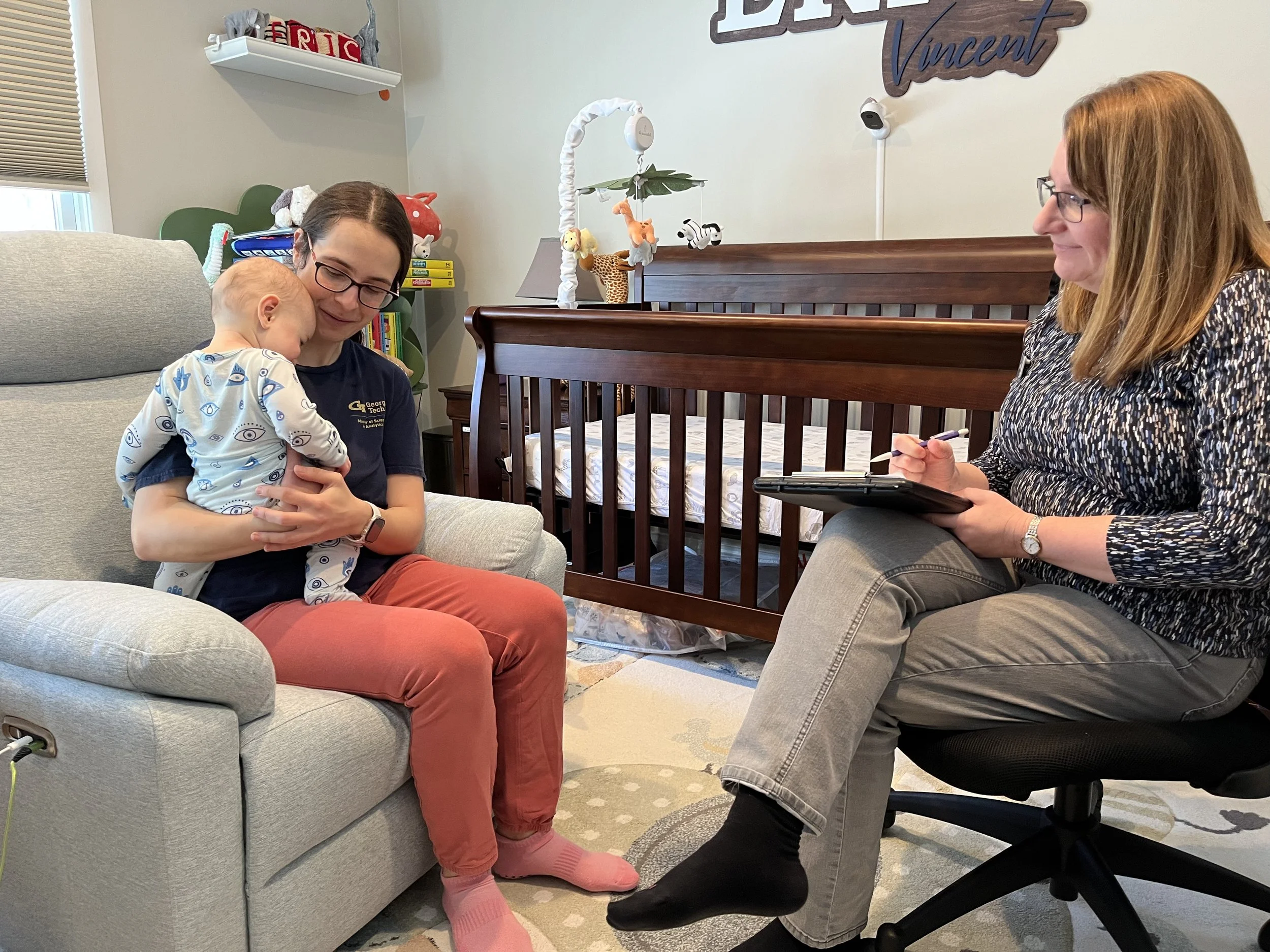 A lactation consultant sitting beside a breastfeeding parent on a couch, gently observing the baby’s latch and offering hands-on support in a cozy living room
