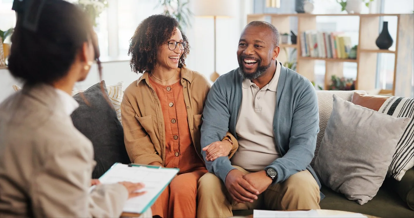 A new parent sitting in a living room holding a sleeping newborn while talking to a therapist on a laptop during a telehealth appointment