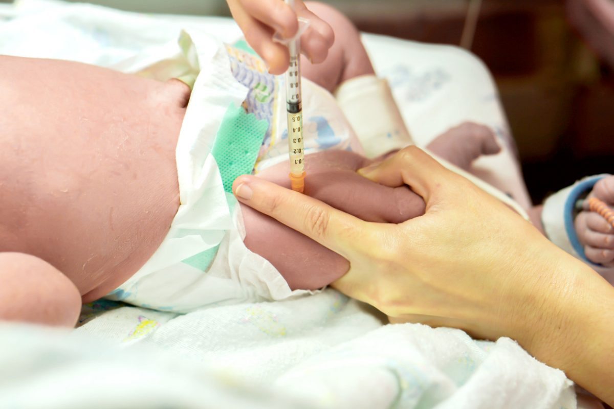 A newborn baby resting on a hospital bassinet mattress while a nurse gently prepares to give a vitamin K injection in the baby’s thigh, candid hospital photography style