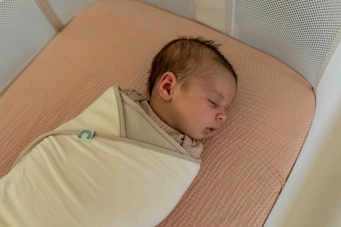 A newborn baby snugly swaddled in a soft cotton swaddle blanket on a bed, arms tucked in, calm neutral lighting, realistic photography