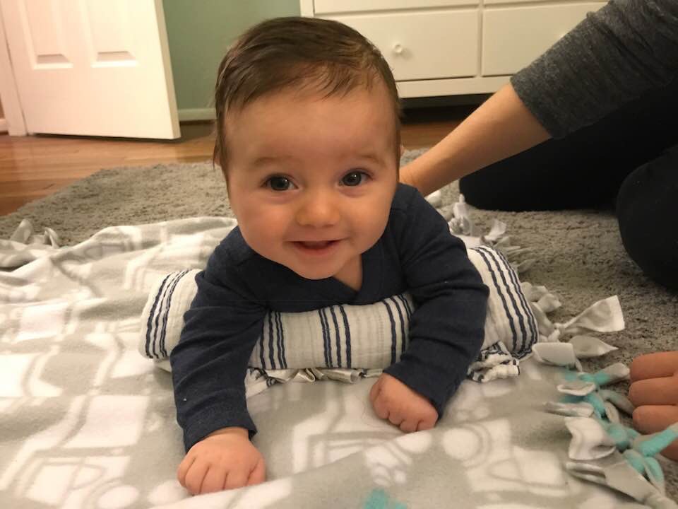 A newborn doing tummy time on a play mat with a small rolled towel placed under their chest for support, a parent sitting nearby with hands close for safety, natural daylight