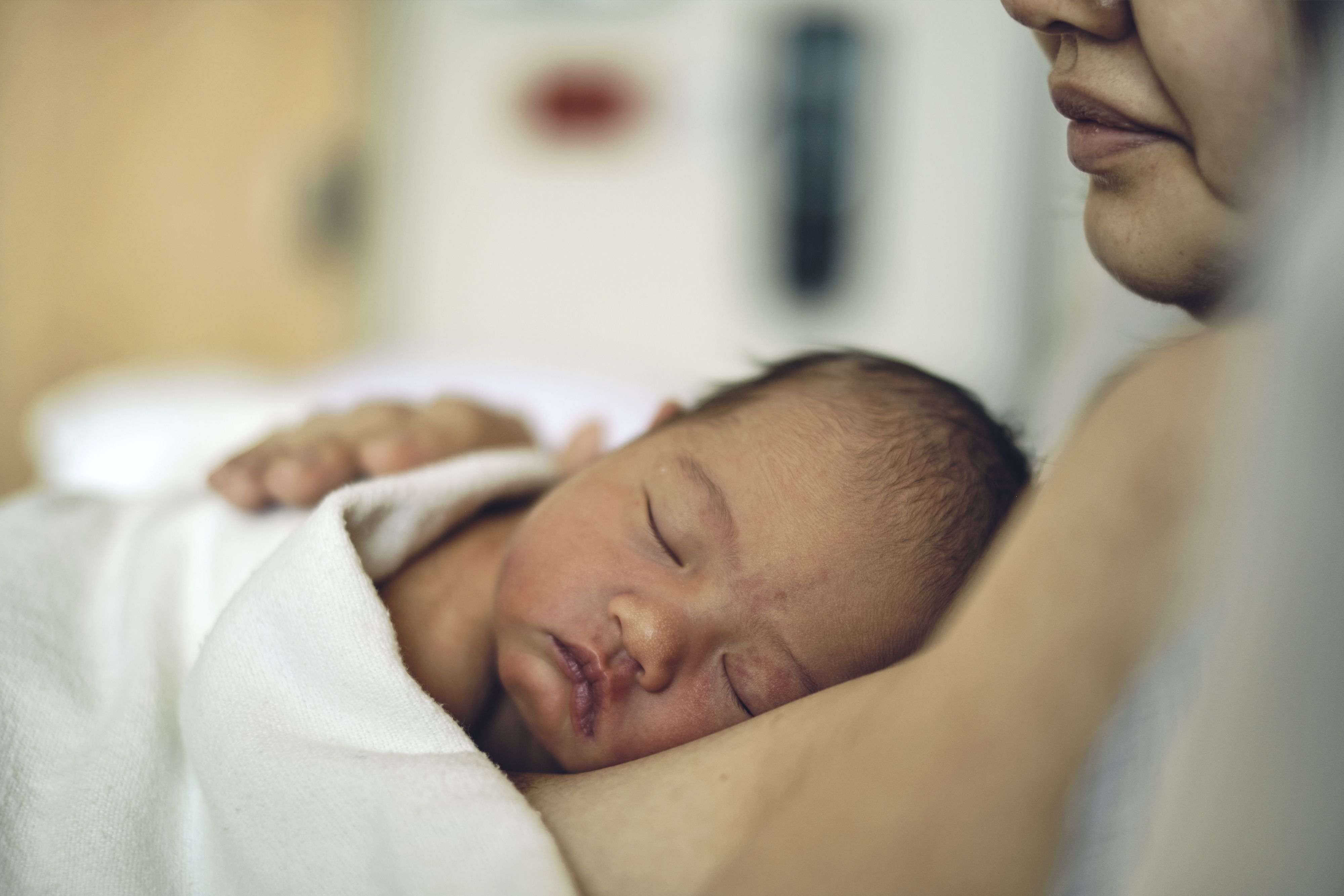 A newborn resting skin-to-skin on a mother's bare chest in a quiet hospital room, both covered with a light blanket