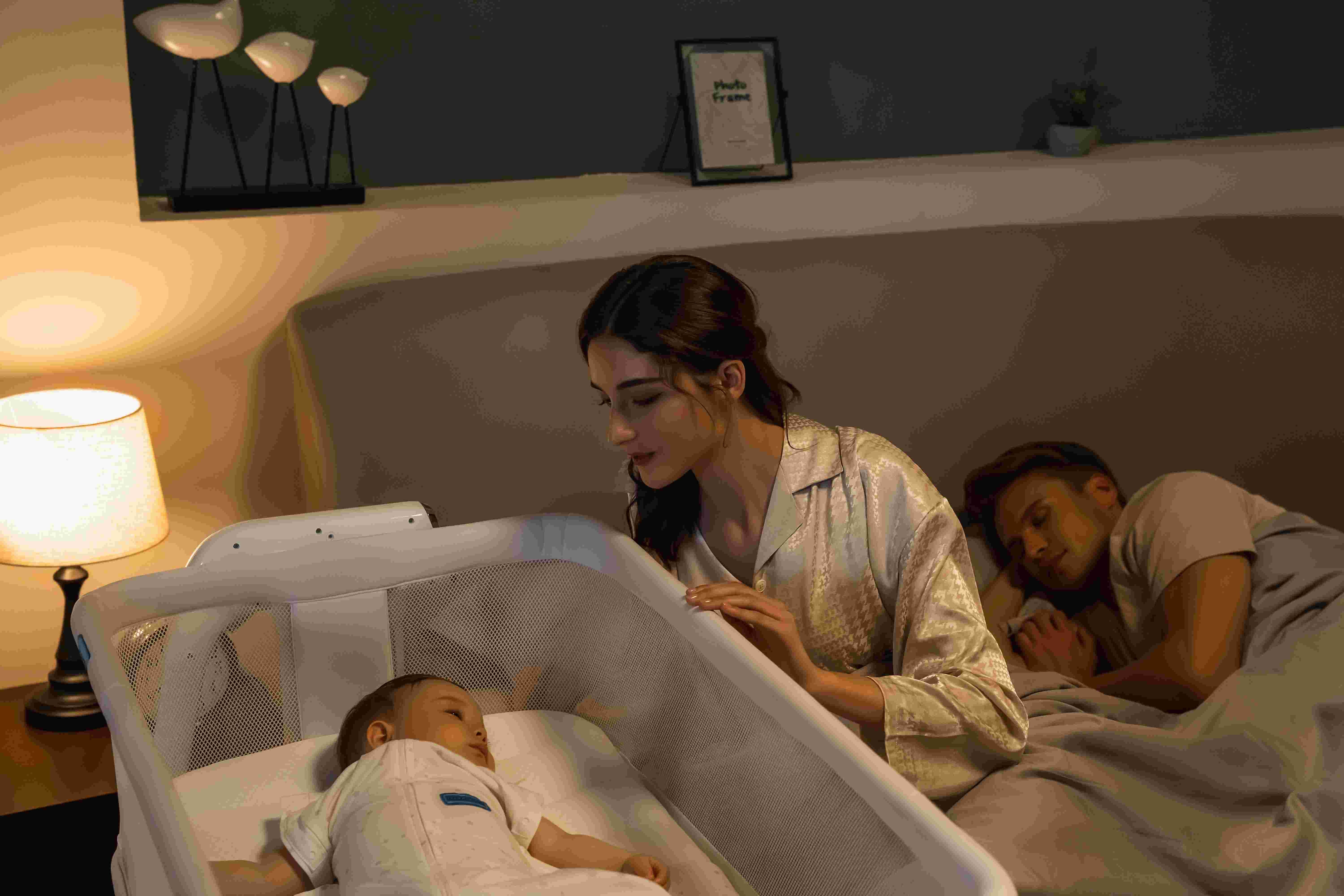 A newborn sleeping on their back in a bedside bassinet next to a neatly made adult bed in a dimly lit bedroom at night, realistic photo