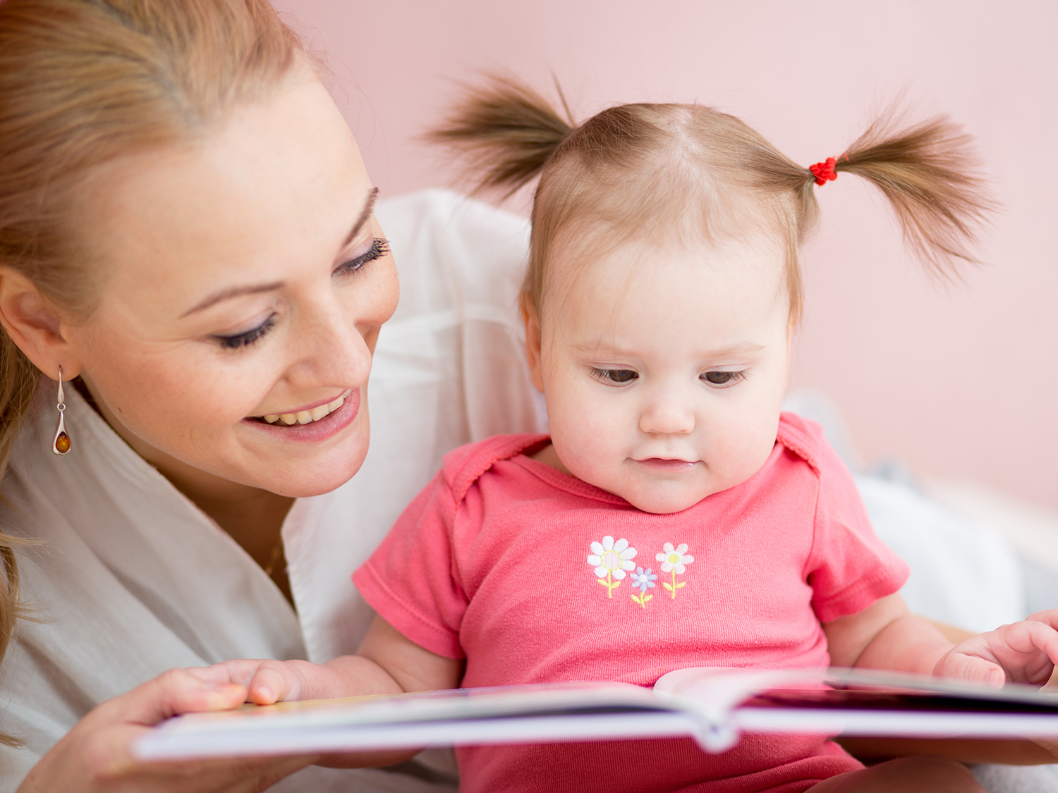 A parent and an 18-month-old toddler sitting on a couch reading a board book together, toddler touching a picture on the page, cozy home setting