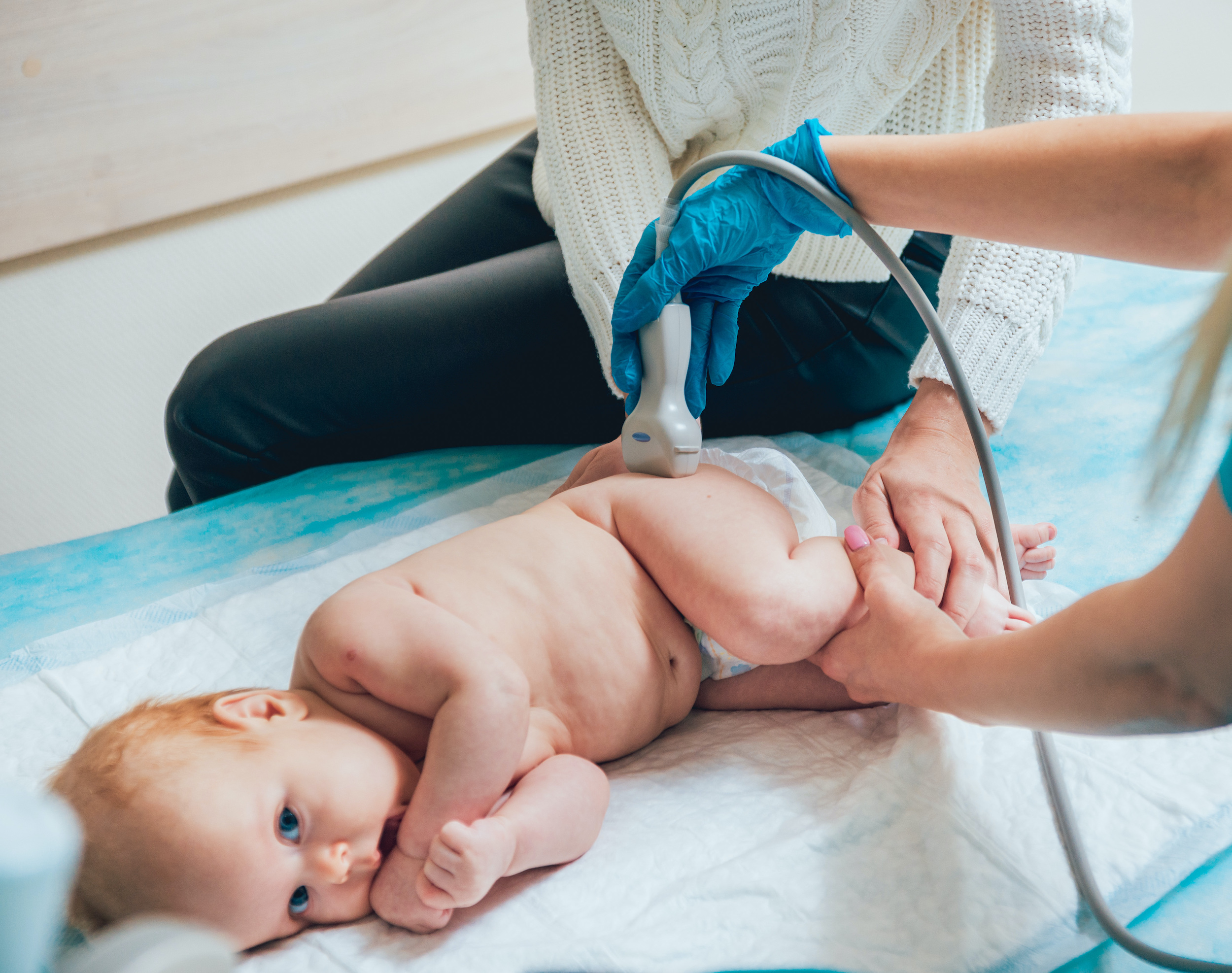 A parent changing a newborn's diaper while a pediatric clinician gently checks the baby's hip and leg position on an exam table in a bright clinic room, realistic photo