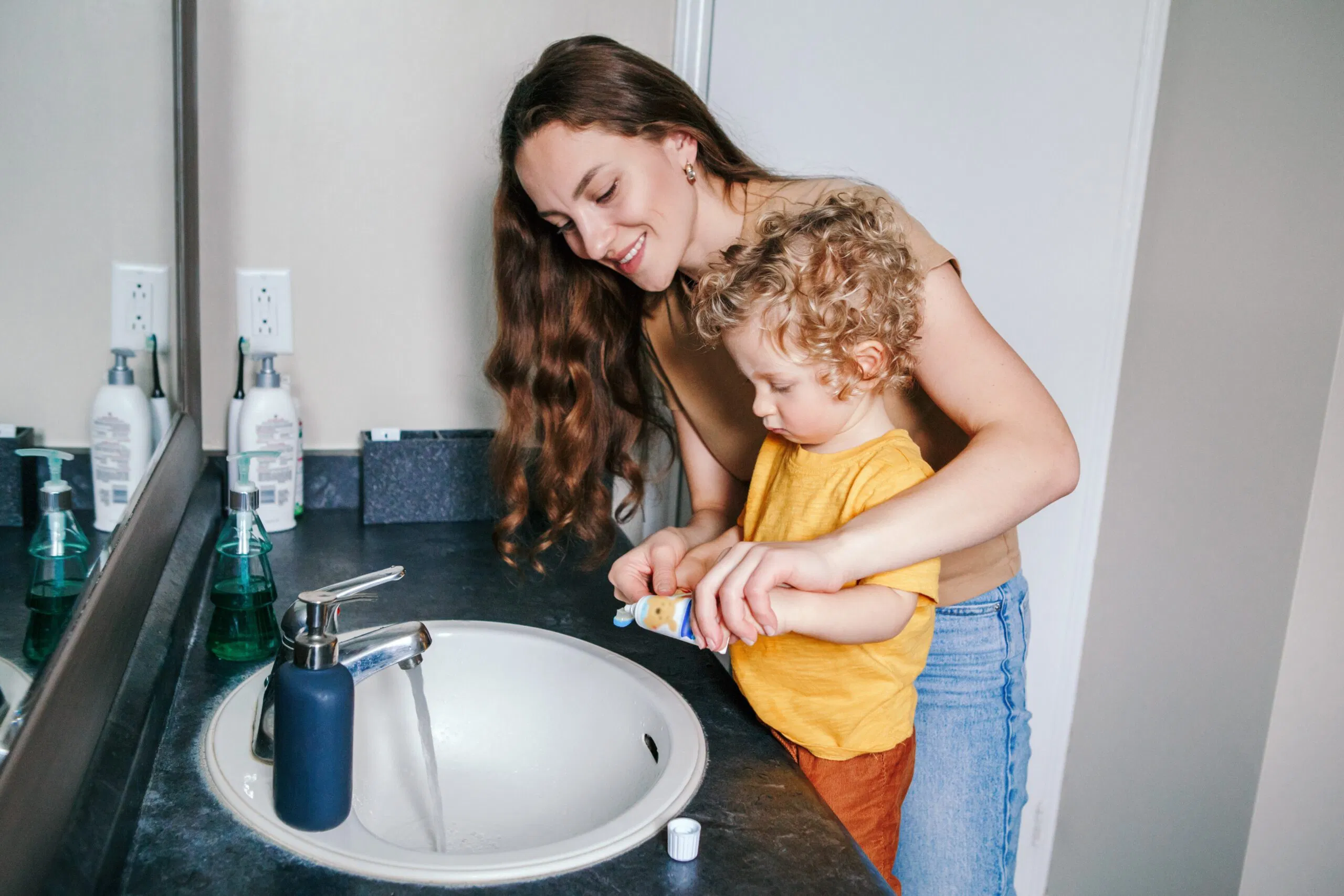 A parent gently brushing a toddler's teeth with a small toothbrush at a bathroom sink during bedtime routine, warm indoor lighting, realistic photo