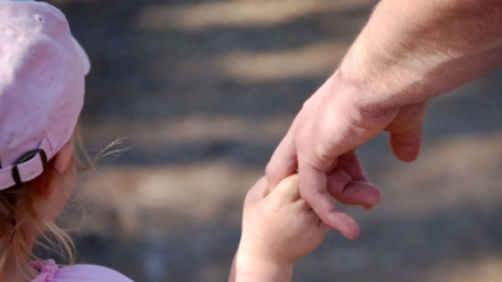 A parent gently holding a toddler’s lower forearm/wrist area while walking through a busy parking lot with cars in the background, realistic documentary photo style