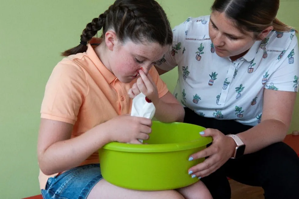 A parent gently pinching the soft part of a toddler's nose while the toddler leans slightly forward on a couch, realistic close-up family photograph