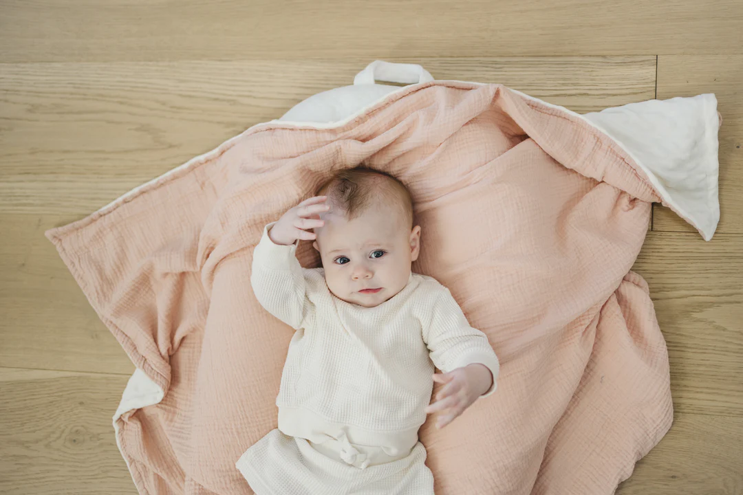 A parent gently tucking a small lightweight toddler blanket over a child lying in a crib, with the blanket positioned at chest level in a calm nursery