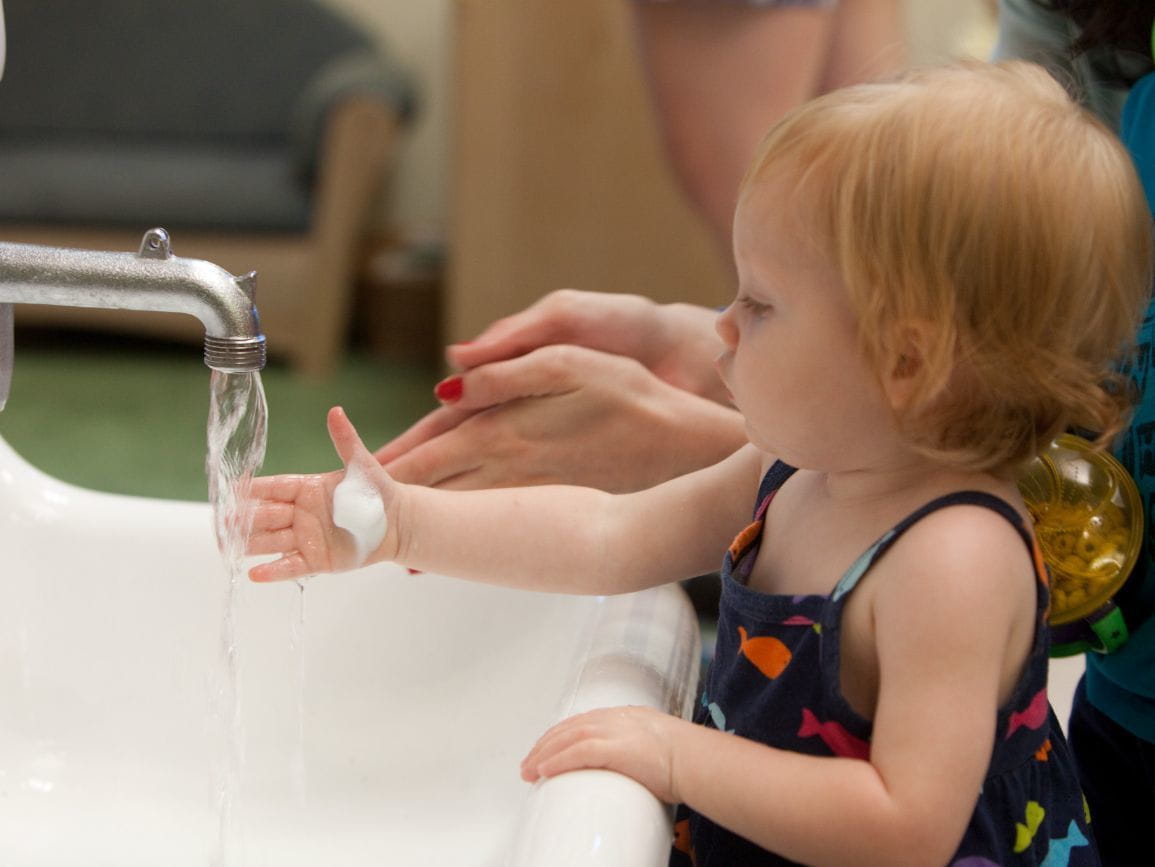 A parent helping a toddler wash hands at a bathroom sink with soap and running water, close-up of hands and faucet, photorealistic candid home photo