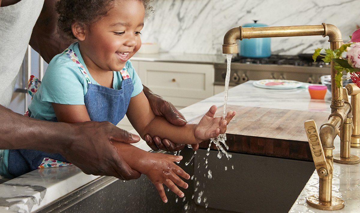 A parent helping a toddler wash hands at a bathroom sink at home, with soap bubbles visible and a calm everyday routine feel