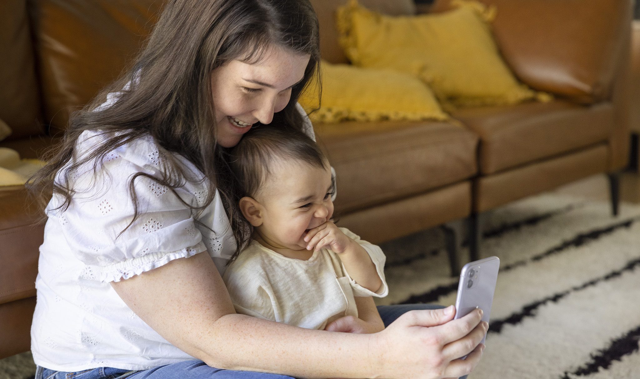 A parent holding a baby on their lap while video chatting with a grandparent on a smartphone, bright daytime living room, natural candid photo