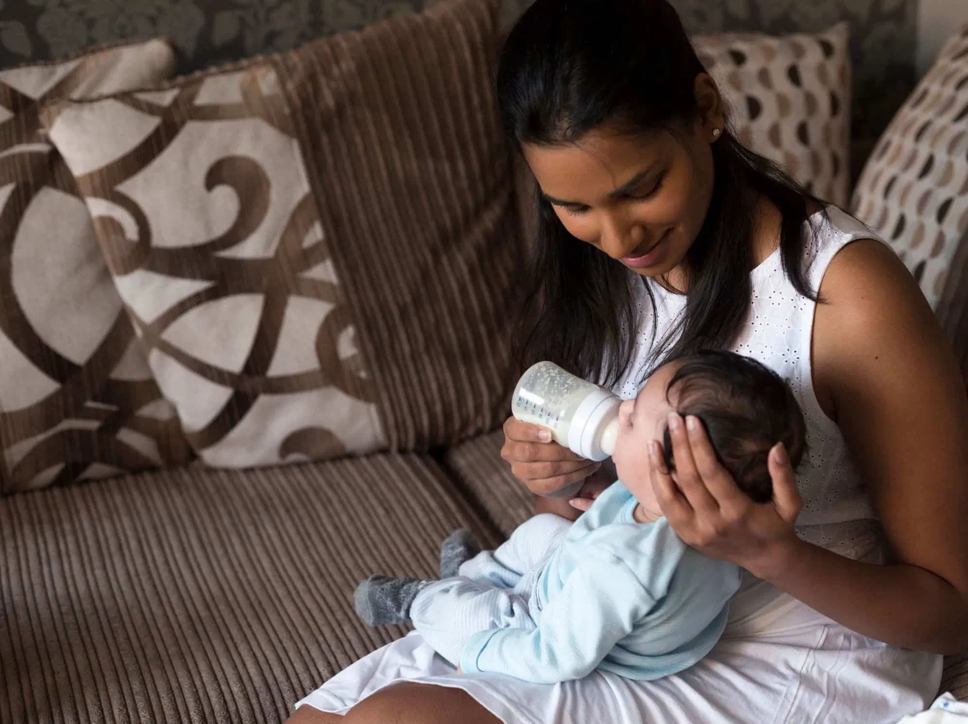 A parent holding a newborn upright in a semi-seated position while offering a bottle slowly, home setting with soft natural light, real photograph