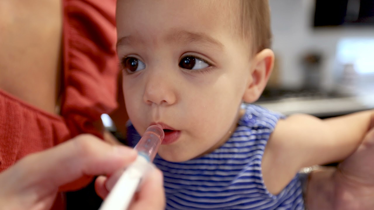 A parent holding a small oral syringe with oral rehydration solution near a baby's mouth while the baby sits supported on a couch, natural home lighting, candid photo