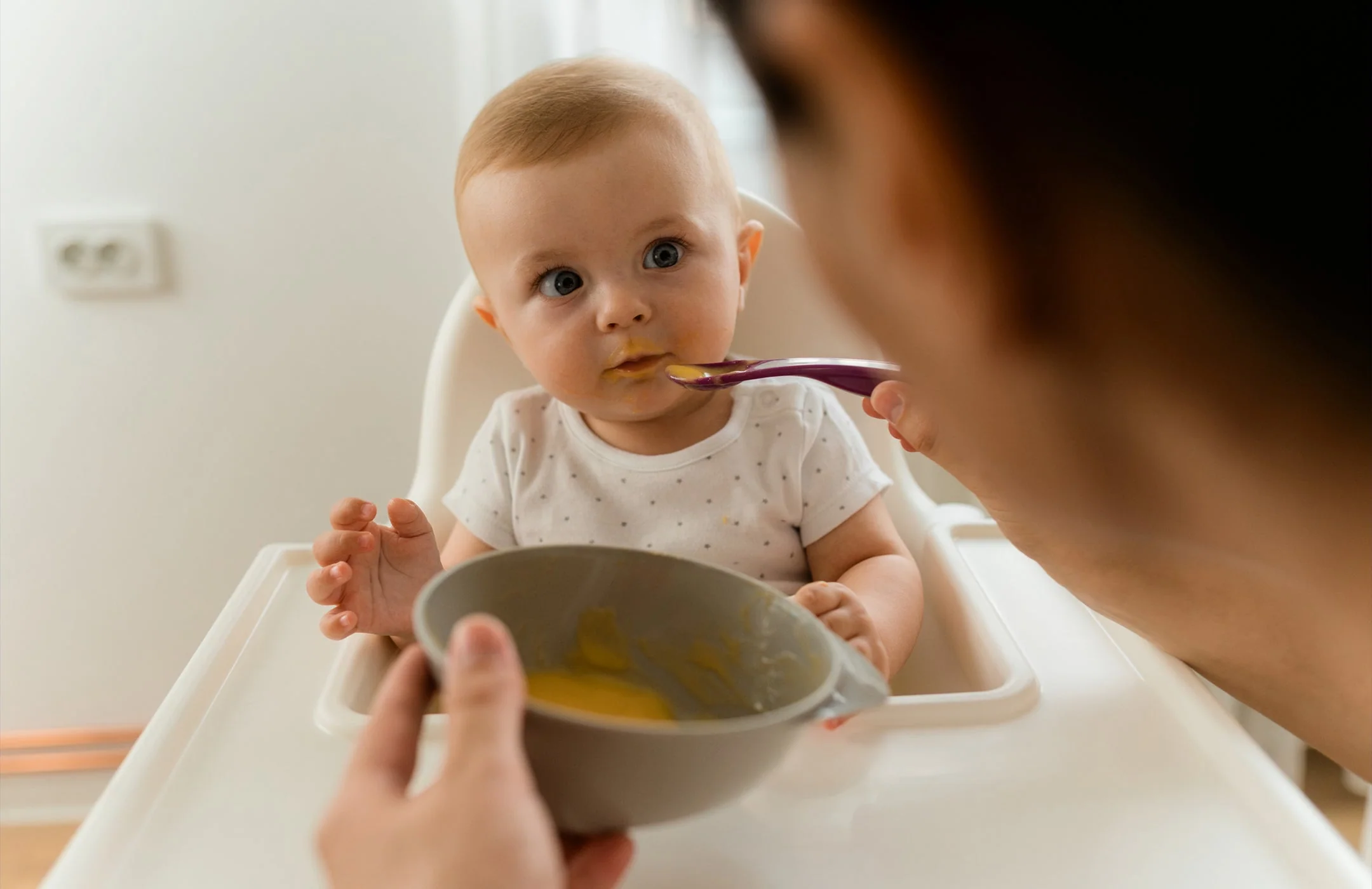 A parent holding a small spoon offering a tiny taste of smooth puree to a 6-month-old baby sitting upright in a high chair, baby leaning forward with curious expression, natural window light, real photograph