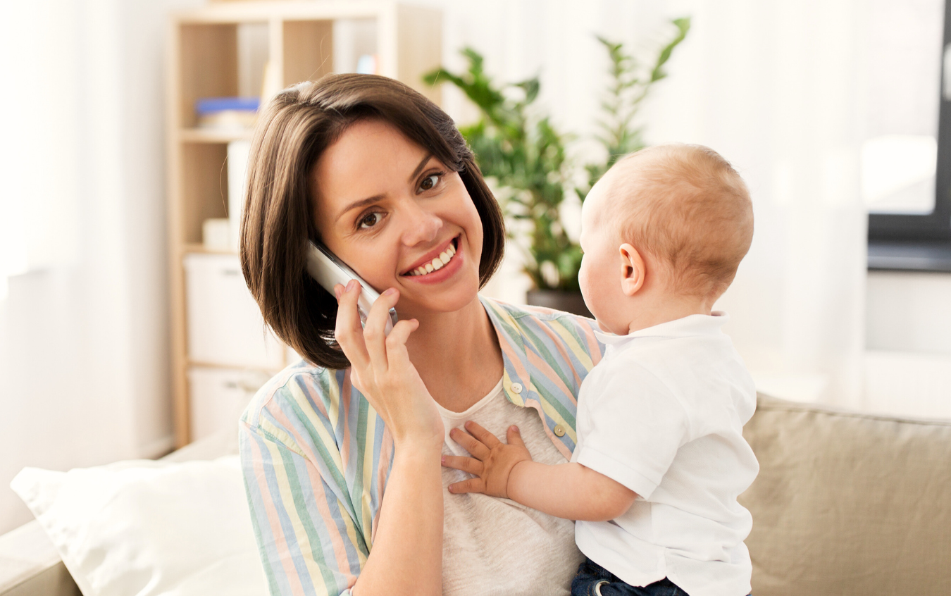 A parent holding a toddler in a pediatric clinic exam room while a clinician reviews test results on a tablet, real-life candid photograph