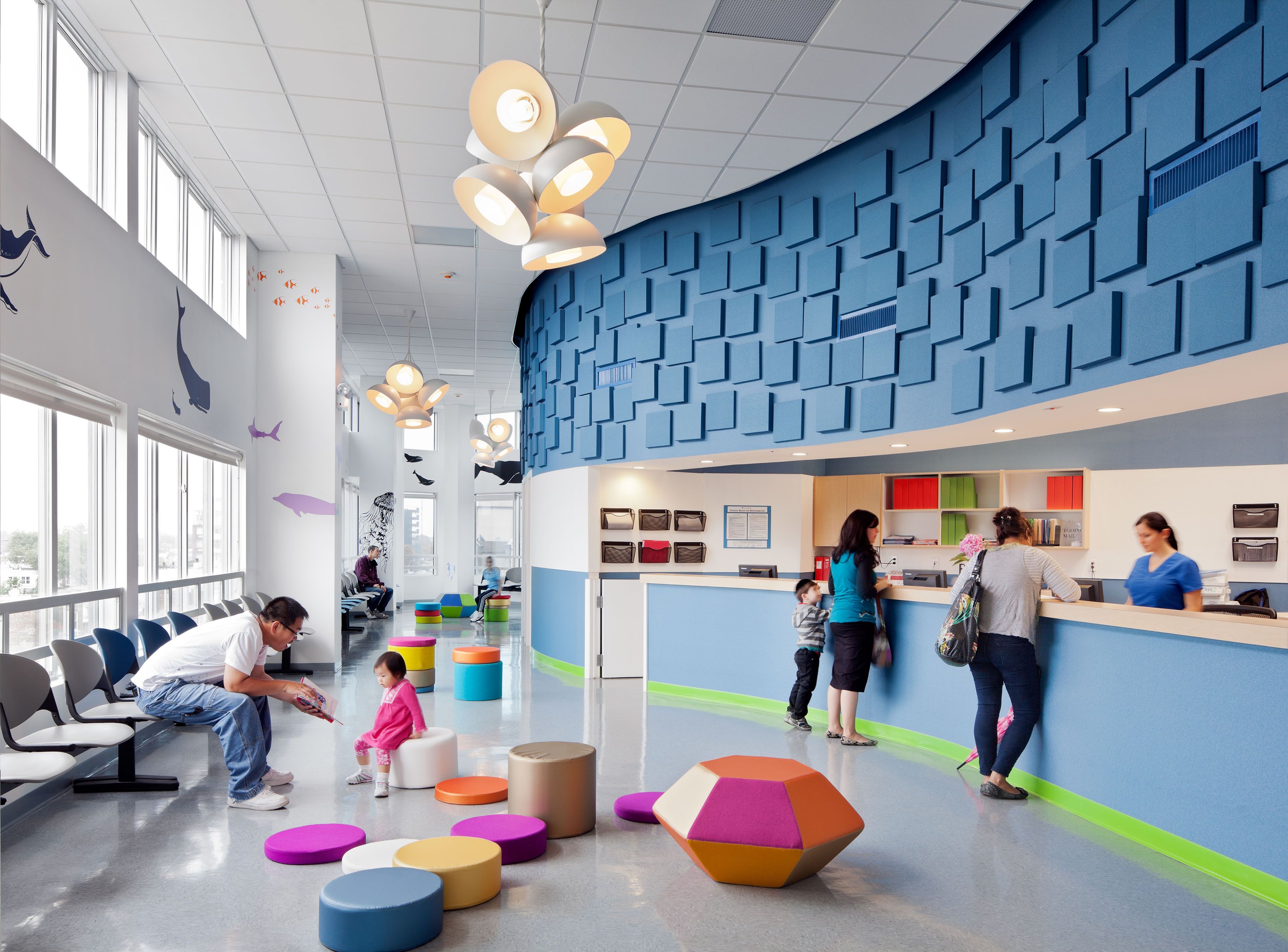 A parent holding a toddler in a pediatric clinic waiting room, looking concerned while reading a phone message, with other families in the background