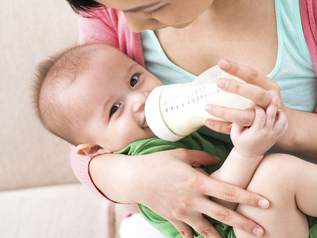 A parent holding an infant who is drinking from a bottle while sitting on a living room couch, natural light, realistic photo