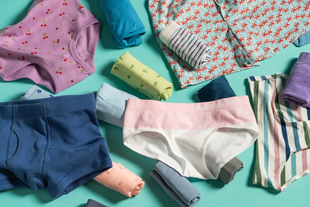 A parent holding clean cotton toddler underwear while a toddler girl stands nearby in a bedroom getting ready for pajamas, natural evening light, real-life photo