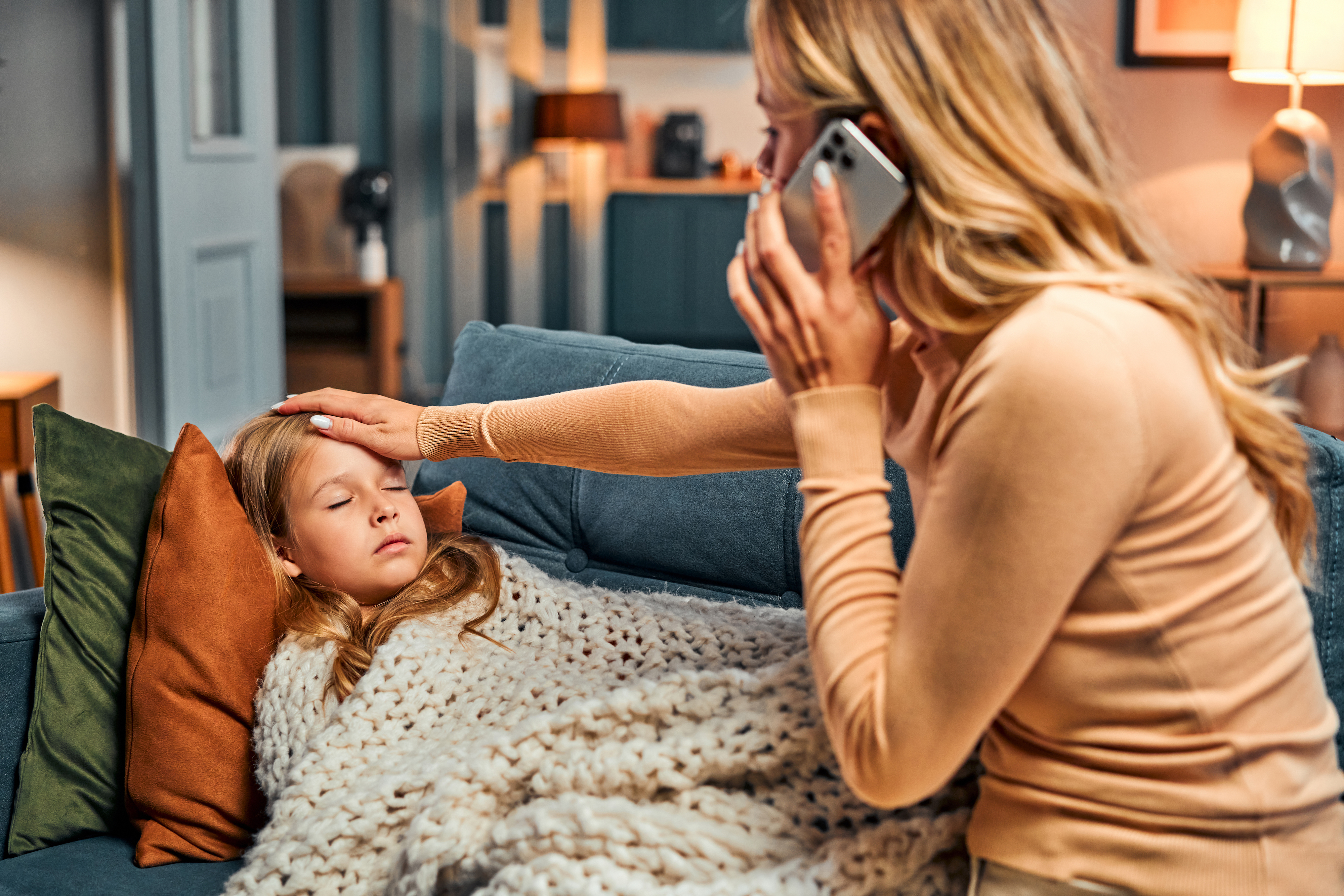 A parent in a kitchen holding a smartphone to their ear while a preschooler plays nearby, capturing the moment of calling a pediatrician for advice