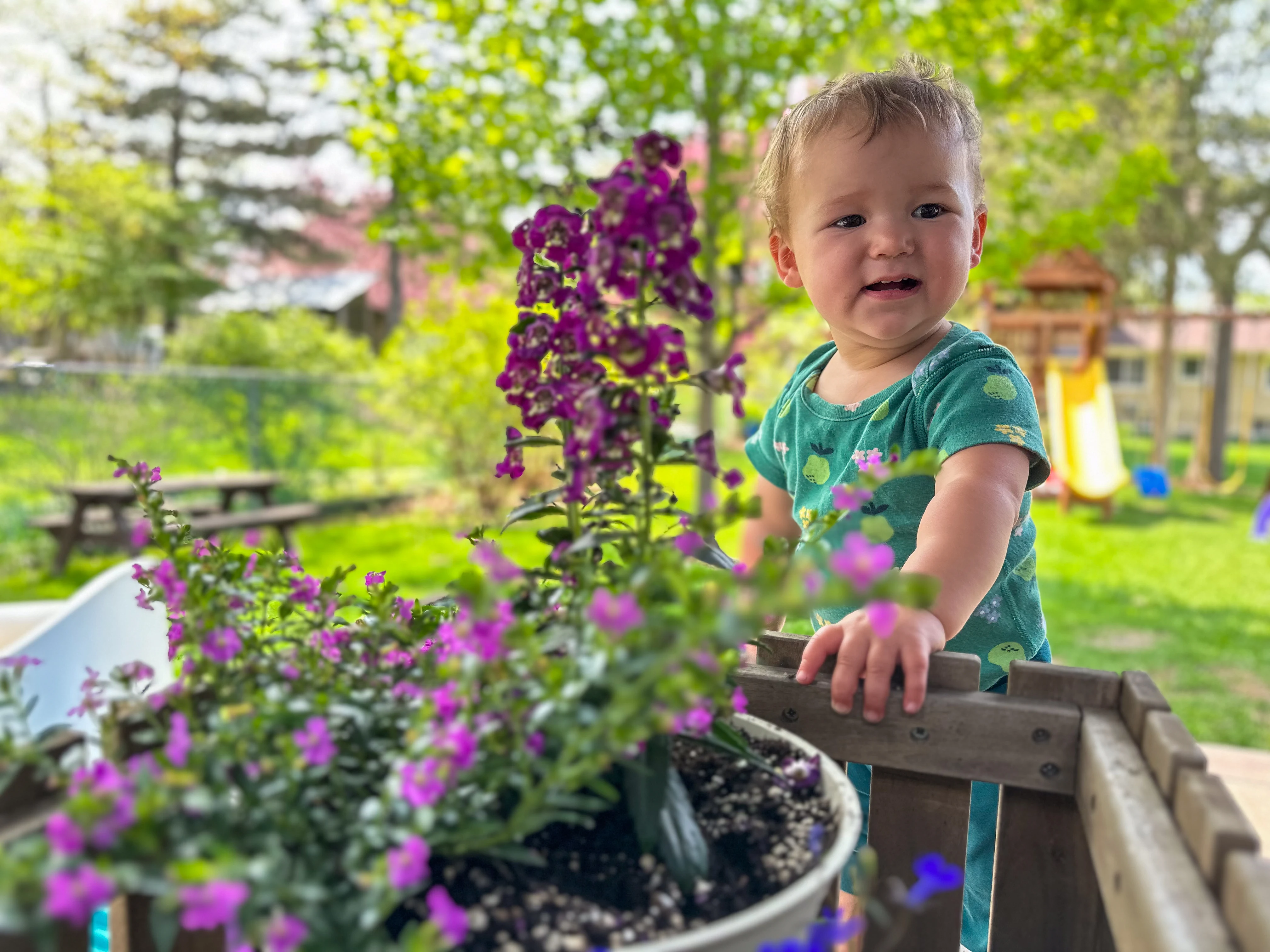 A parent kneeling at a playground gently holding a toddler’s hand away from a patch of dirt while offering a snack, candid outdoor photo