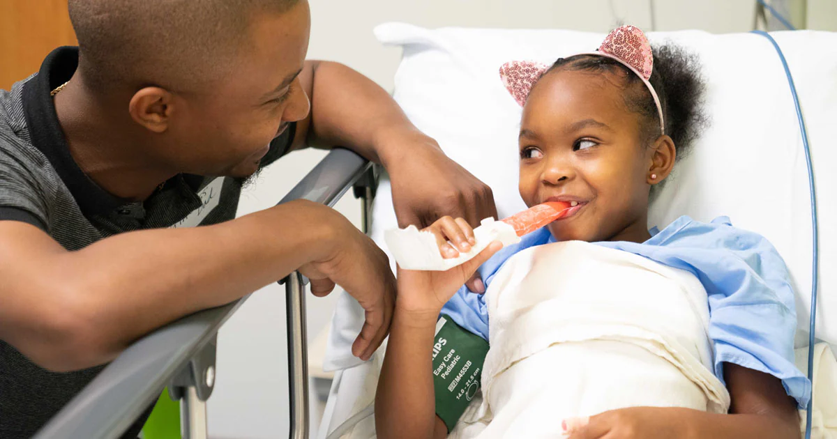 A parent kneeling beside a couch offering a popsicle to a sick toddler wrapped in a blanket, soft indoor light