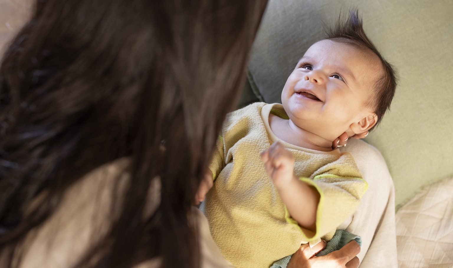 A parent leaning over a baby on a couch while the baby smiles and makes eye contact, soft indoor light, candid family photo