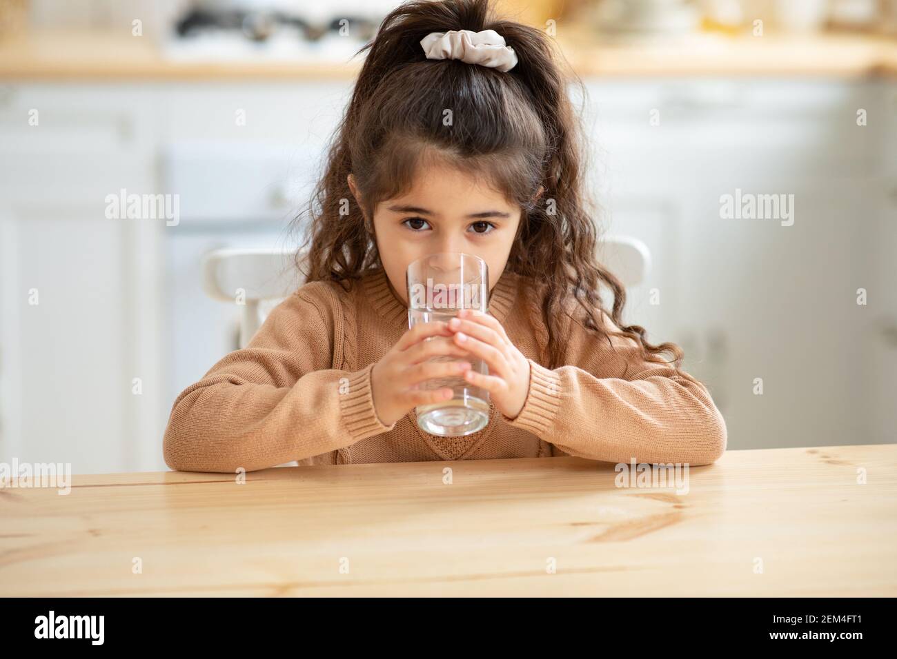 A parent offering a glass of water to a school-aged child sitting at a kitchen table with a blanket around their shoulders, candid home photo