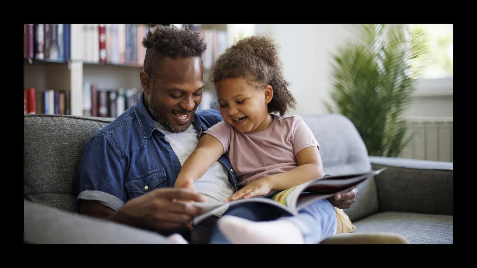 A parent reading a picture book on a couch while a toddler looks at the pages, natural indoor photo