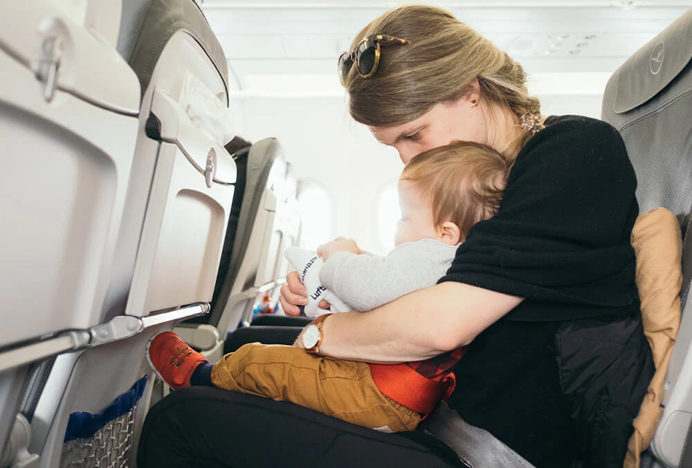 A parent seated on an airplane holding a baby and offering a bottle near the window, candid travel photo