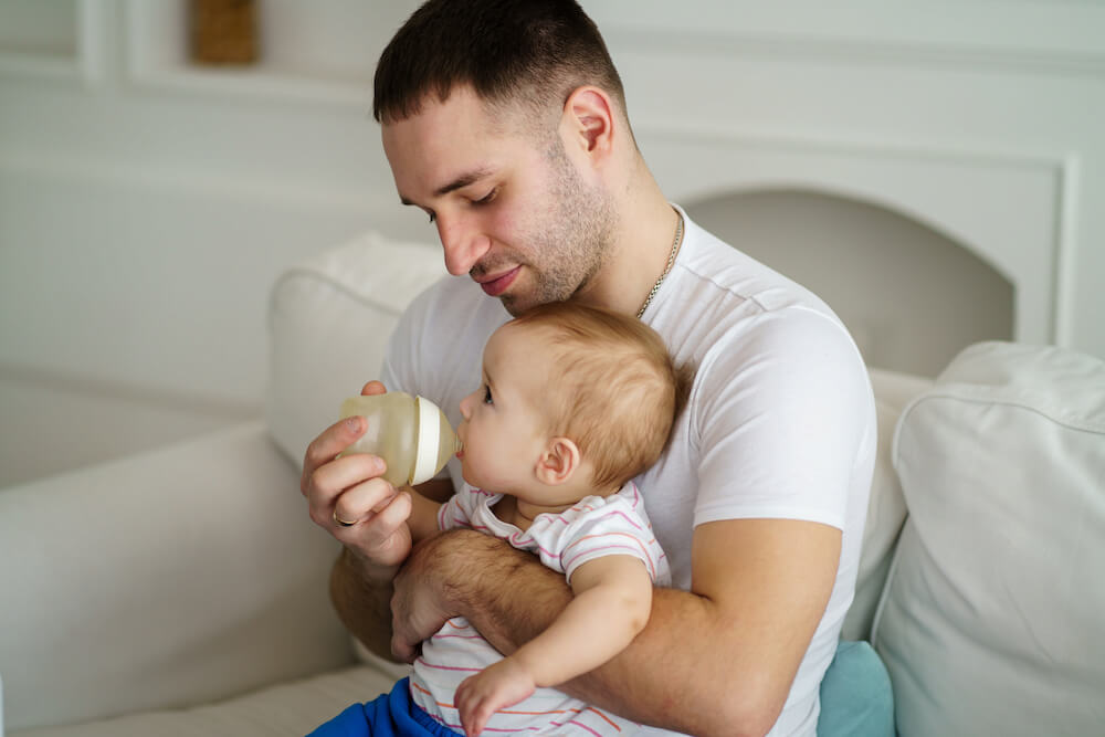 A parent sitting on a couch feeding an infant a bottle in an upright, supported position with the baby calm and relaxed, natural indoor light, realistic photo