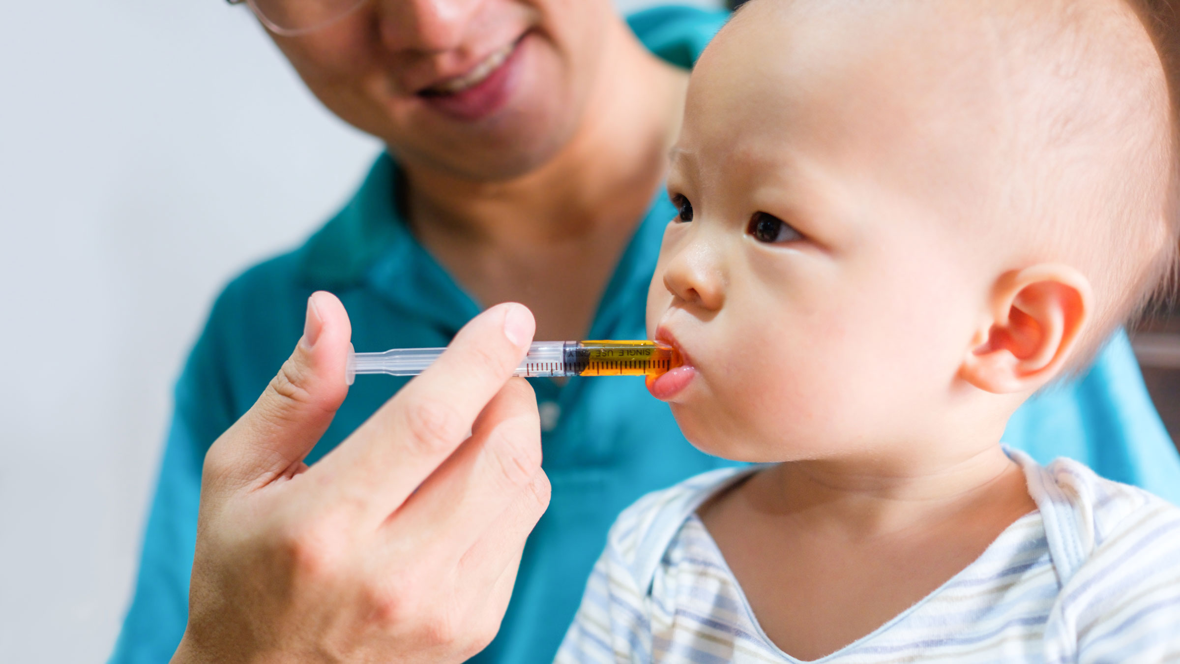 A parent sitting on a couch gently giving a toddler liquid medicine with an oral syringe, the toddler seated upright on the parent’s lap in a calm living room, natural window light, candid family photo
