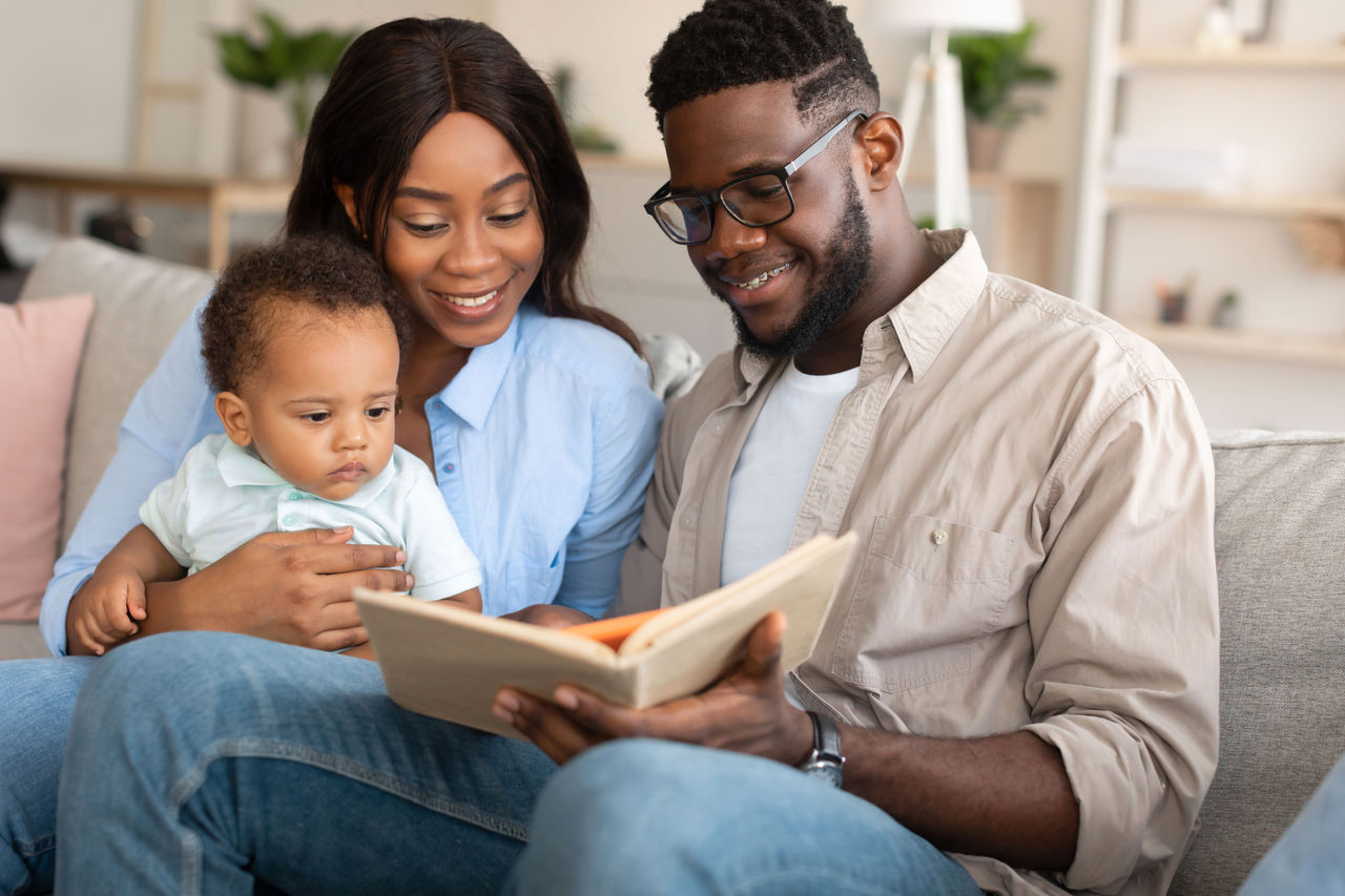 A parent sitting on a couch reading a picture book with a toddler who points at the pages, cozy home setting with warm lighting