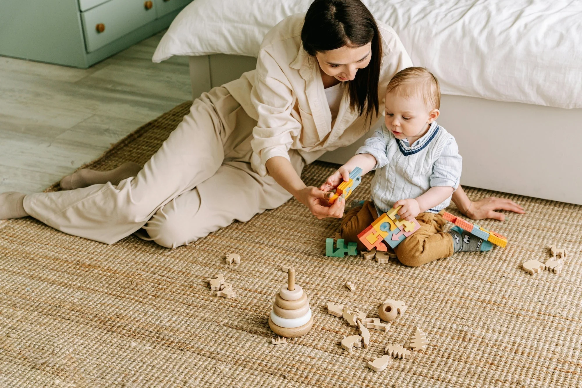 A parent sitting on the floor while a baby reaches forward toward a toy on a soft rug in a cozy living room, natural candid family photo
