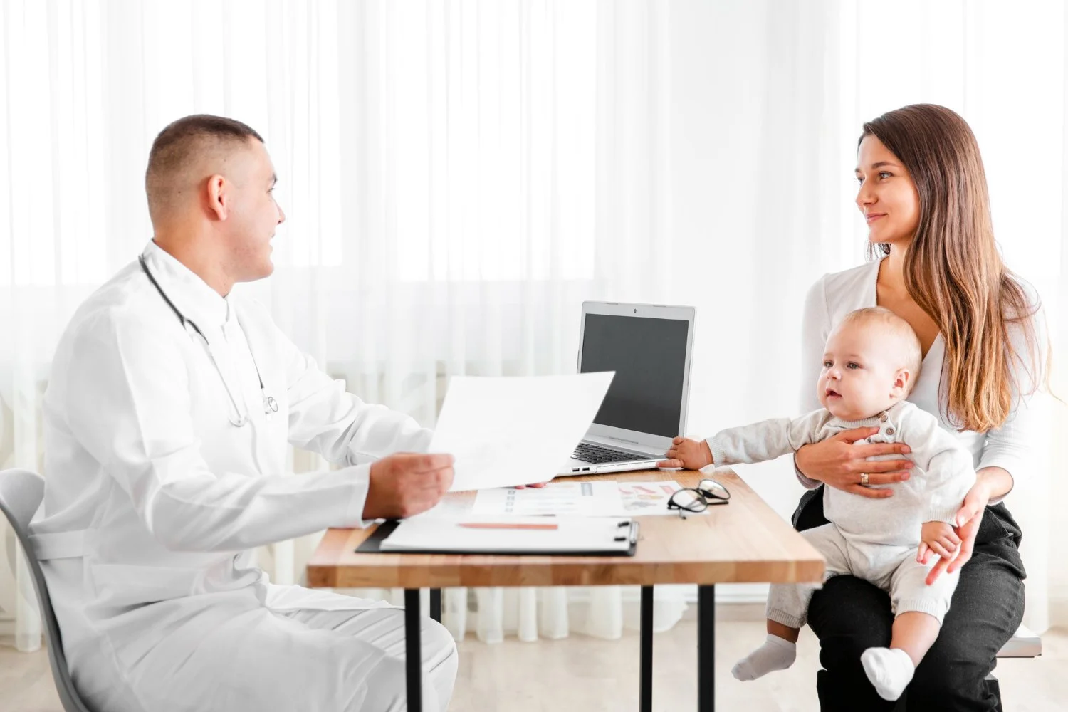 A parent sitting with a pediatrician in a clinic exam room while holding a preschooler on their lap, realistic medical visit photo