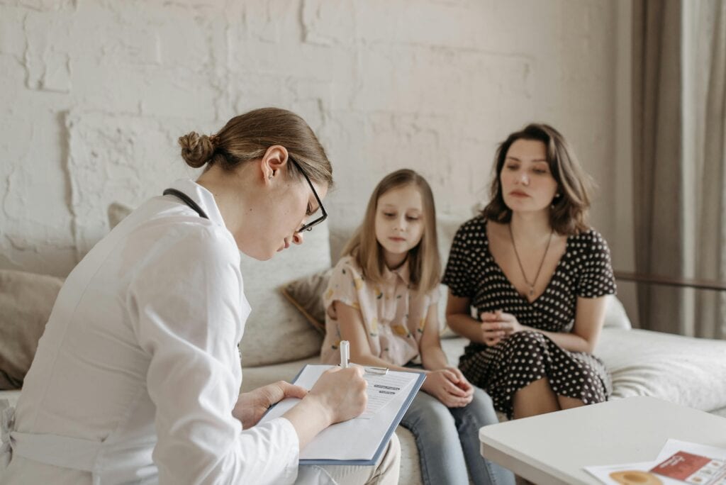 A parent sitting with a toddler on their lap while speaking with a pediatrician in a bright exam room, realistic medical office photograph
