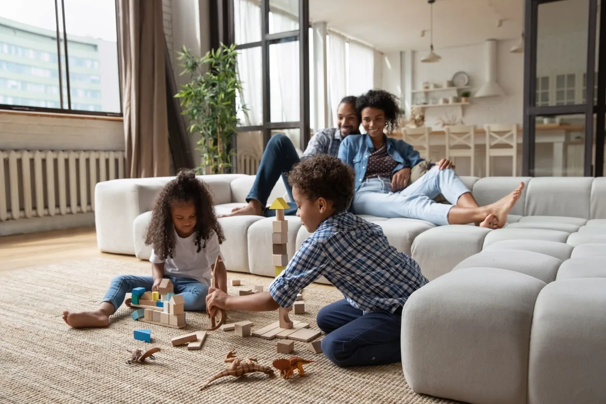 A parent vacuuming a living room rug with a modern vacuum while a toddler plays with blocks nearby, candid home photo